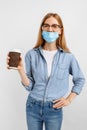 Beautiful young woman in a medical protective mask on her face holding a paper Cup of coffee on a white background