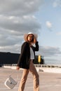 Beautiful young woman model in straw hat in black blazer in fashionable pants in casual sneakers with bag walk from store. Royalty Free Stock Photo