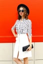 Beautiful young woman model wearing white striped shirt, black handbag clutch, round hat, skirt on city street over red wall Royalty Free Stock Photo