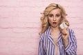 Beautiful young woman in pajamas standing isolated on pink background picking up chocolate and being surprised.