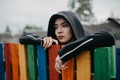 Young woman in a hood with a pensive look stands by a multicolored rainbow fence