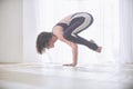 Beautiful young woman practices yoga asana bakasana - crane pose in the light yoga studio