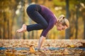 Beautiful young woman practices yoga asana Bakasana - crane pose on the wooden deck in the autumn park.