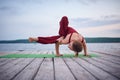 Beautiful young woman practices yoga asana Parivritta Eka Pada Danda Kaundiniasana on the wooden deck near the lake