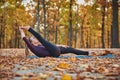 Beautiful young woman practices yoga asana Supta Padangushthasana 1 on the wooden deck in the autumn park.