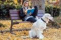 Beautiful young woman reading a book while sitting on bench with her lovely golden retriever dog in the park in autumn Royalty Free Stock Photo