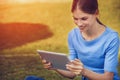 A beautiful young woman is reading from a tablet pc while seating outdoors on grass in a park Royalty Free Stock Photo
