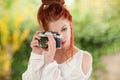 Beautiful young woman with red hair sitting in the garden taking pictures with camera Royalty Free Stock Photo