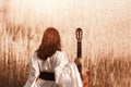 Beautiful young woman with red hair in a white medieval dress holding guitar and walking through the sunny field at sunset Royalty Free Stock Photo