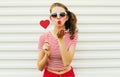 Beautiful young woman with red heart shaped sunglasses blowing lips sending sweet air kiss on white wall Royalty Free Stock Photo
