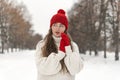 Beautiful young woman in red knitted hat and mittens and woolen sweater on alley in winter park. Portrait of pretty girl