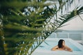 Beautiful young woman relaxing in pool under tropical palm leaves, enjoying day. Brunette girl on vacation in luxury resort, Royalty Free Stock Photo