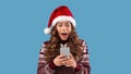Beautiful young woman in Santa hat with smartphone shocked over big Christmas sale in online store on blue background