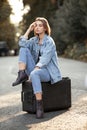 Beautiful young woman seated on her suitcase on middle road, waiting a transportation, in sunset background Royalty Free Stock Photo