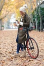 Beautiful young woman sending messages with her smart phone while cycling through the park in autumn Royalty Free Stock Photo