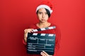 Beautiful young woman with short hair wearing christmas hat holding clapboard puffing cheeks with funny face Royalty Free Stock Photo