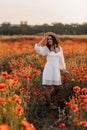 Beautiful young woman in short white dress and straw hat in field with poppies in evening at sunset and holds poppy in hand, Royalty Free Stock Photo