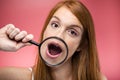 Beautiful young woman showing her mouth trought magnifying glass over pink background.