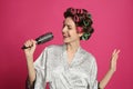 Beautiful young woman in silk bathrobe with hair curlers singing into hairbrush on pink background Royalty Free Stock Photo