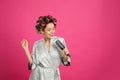 Beautiful young woman in silk bathrobe with hair curlers singing into hairbrush on pink background. Space for text Royalty Free Stock Photo