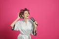 Beautiful young woman in silk bathrobe with hair curlers singing into hairbrush on pink background. Space for text Royalty Free Stock Photo