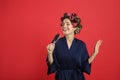 Beautiful young woman in silk bathrobe with hair curlers singing into hairbrush on red background Royalty Free Stock Photo