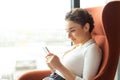 Beautiful young woman sitting in an armchair in a hotel room by the window and using her smart phone Royalty Free Stock Photo