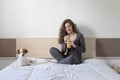 beautiful young woman sitting on bed with her cute small dog besides. Home, indoors and lifestyle. She is drinking orange juice.