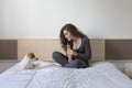 beautiful young woman sitting on bed with her cute small dog besides. Home, indoors and lifestyle. She is drinking orange juice.