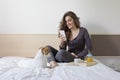 Beautiful young woman sitting on bed with her cute small dog besides. Home, indoors and lifestyle. She is having breakfast on bed Royalty Free Stock Photo