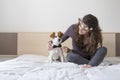 beautiful young woman sitting on bed with her cute small dog besides. Dog wearing bowtie and the girl is wearing a flowers wreath