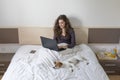 Beautiful young woman sitting on bed with her cute small dog besides. She is working on laptop and smiling. Breakfast at home, Royalty Free Stock Photo