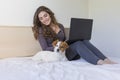 beautiful young woman sitting on bed with her cute small dog besides. She is working on laptop and smiling. Breakfast at home, Royalty Free Stock Photo