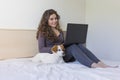 beautiful young woman sitting on bed with her cute small dog besides. She is working on laptop and smiling. Breakfast at home,