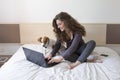 Beautiful young woman sitting on bed with her cute small dog besides. She is working on laptop and smiling. Home, indoors and Royalty Free Stock Photo