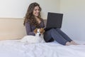 Beautiful young woman sitting on bed with her cute small dog besides. She is working on laptop and smiling. Home, indoors and Royalty Free Stock Photo