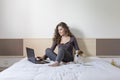 Beautiful young woman sitting on bed with her cute small dog besides. She is working on laptop and smiling. Home, indoors and Royalty Free Stock Photo