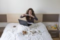 Beautiful young woman sitting on bed with her cute small dog besides. She is working on laptop and taking a picture of her dog Royalty Free Stock Photo