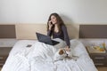 beautiful young woman sitting on bed with her cute small dog besides. She is working on laptop and talking on her mobile phone. Royalty Free Stock Photo