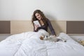 Beautiful young woman sitting on bed with her cute small dog besides. She is working on tablet and smiling. Breakfast at home, Royalty Free Stock Photo