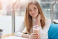 Beautiful young woman sitting in cafe with fresh cocktail, having rest after lectures, looks smiling at camere, keeps hand on neck Royalty Free Stock Photo