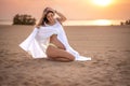 Beautiful young woman sitting on sand, holding her white dress and enjoying being pregnant on the beach at sunset. Royalty Free Stock Photo