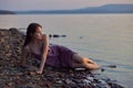 Beautiful young woman sitting on shore of lake in summer dress at sunset. Portrait of a romantic wet girl at sunset, warm sun, Royalty Free Stock Photo