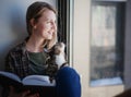 Beautiful young woman sitting on a window sill with a striped cat and reading a book Royalty Free Stock Photo
