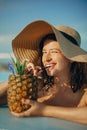 Beautiful young woman smiling and drinking cocktail in pineapple, relaxing in pool on summer vacation. Girl in hat enjoying warm Royalty Free Stock Photo