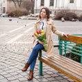 Beautiful young woman with spring tulips flowers bouquet at city street. Happy girl sitting on a bench outdoors. Spring full body Royalty Free Stock Photo