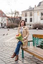 Beautiful young woman with spring tulips flowers bouquet at city street. Happy girl sitting on a bench outdoors. Spring full body Royalty Free Stock Photo