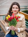 Beautiful young woman with spring tulips flowers bouquet at city street. Happy girl sitting on a bench outdoors. Spring portrait Royalty Free Stock Photo