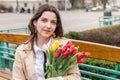 Beautiful young woman with spring tulips flowers bouquet at city street. Happy girl sitting on a bench outdoors. Spring portrait Royalty Free Stock Photo