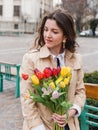 Beautiful young woman with spring tulips flowers bouquet at city street. Happy girl sitting on a bench outdoors. Spring portrait Royalty Free Stock Photo